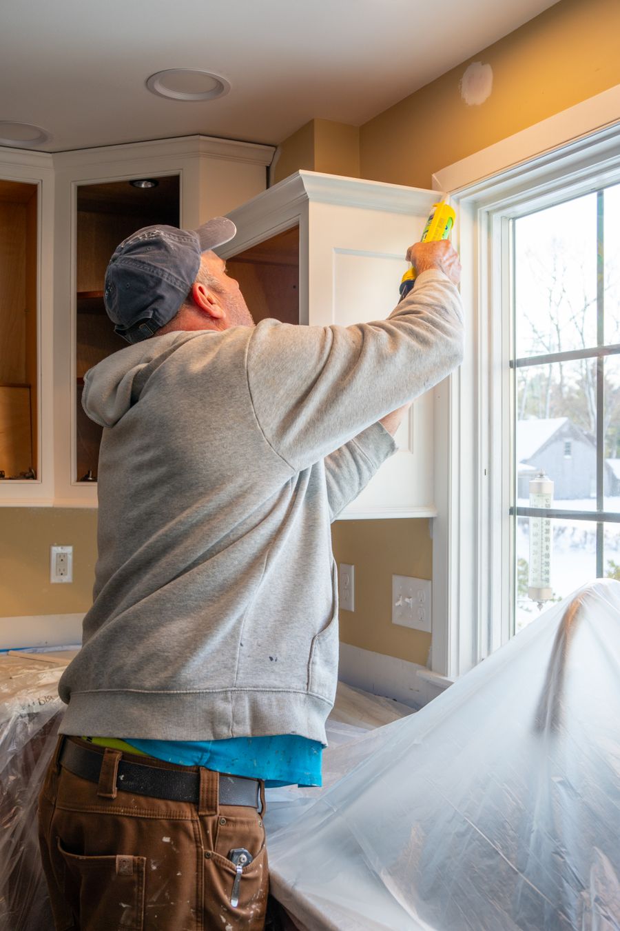 Technician running a fresh bead of caulk along kitchen cabinet trim beside a window
